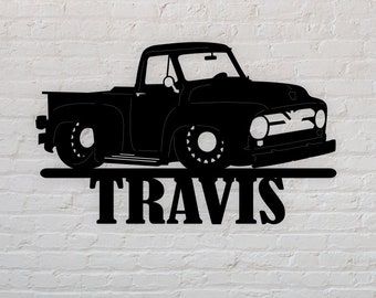 Op de afbeelding: Zwart metalen bord met een silhouet van een klassieke pick-up truck. De truck is gedetailleerd met wielen, een cabine en een laadbak. De naam "TRAVIS" staat in vette letters eronder, voor interieurdecoratie.