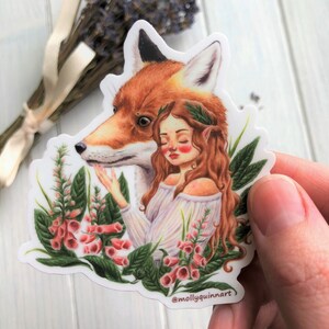 May include: A sticker featuring a woman with long red hair and a fox head. She is wearing a white dress and has a crown of leaves on her head. The sticker is surrounded by pink flowers and green leaves. The text "@mollyquinnart" is at the bottom of the sticker.