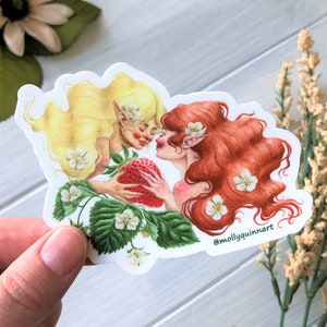 May include: A sticker featuring two fairies with long flowing hair, one with blonde hair and the other with red hair. They are holding a large red strawberry between them and are looking at each other. The sticker has a white background and is surrounded by green leaves and white flowers. The sticker has the text "@mollyquinart" in the bottom right corner.