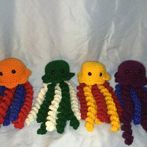 May include: Four crocheted jellyfish toys in different colors. The jellyfish are orange, green, yellow, and purple. Each jellyfish has a white crocheted belly and a curled tentacle.