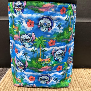 May include: A cylindrical fabric container with a tropical design featuring the cartoon character Stitch. The pattern includes palm trees, red hibiscus flowers, and Stitch against a blue sky. The top edge of the container is black.