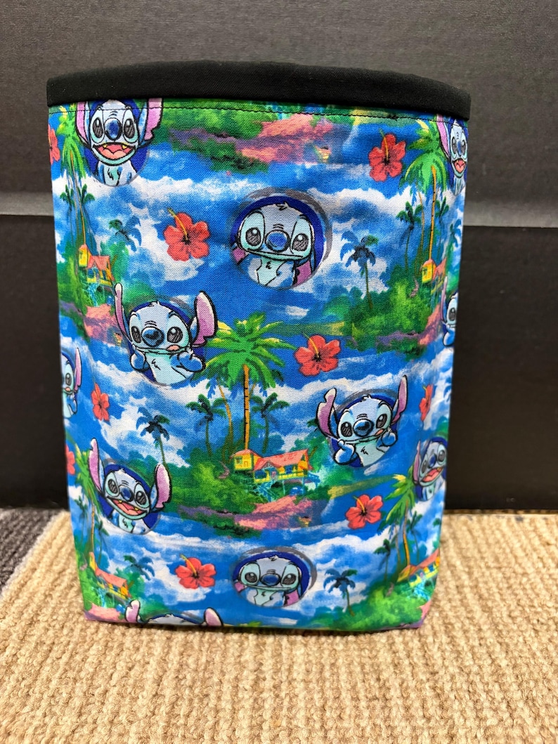 May include: A cylindrical fabric container with a tropical design featuring the cartoon character Stitch. The pattern includes palm trees, red hibiscus flowers, and Stitch against a blue sky. The top edge of the container is black.