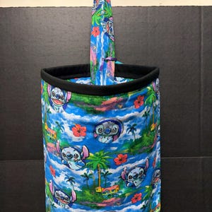May include: A cylindrical fabric container with a black rim and a tall, pointed, surfboard-shaped insert. The container and insert feature a vibrant print of Stitch from Lilo & Stitch, palm trees, and tropical flowers against a blue sky.