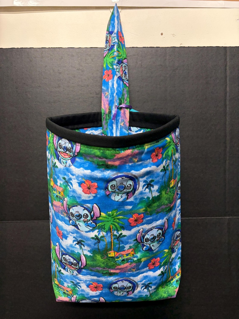May include: A cylindrical fabric container with a black rim and a tall, pointed, surfboard-shaped insert. The container and insert feature a vibrant print of Stitch from Lilo & Stitch, palm trees, and tropical flowers against a blue sky.