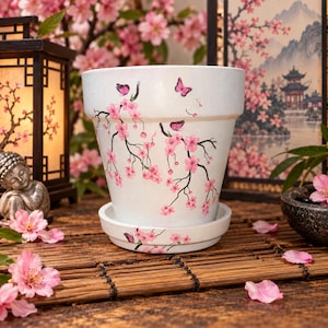 May include: A white ceramic plant pot with a matching saucer, decorated with hand-painted black branches, pink cherry blossoms, and small pink butterflies. The design wraps around the exterior of the pot and continues onto the rim of the saucer.