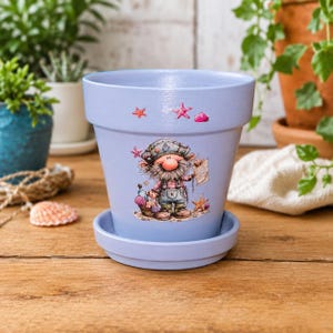 May include: A light blue ceramic flower pot with a matching saucer, featuring a whimsical illustration of a bearded gnome wearing a sailor hat and overalls, surrounded by seashells and starfish, with additional small starfish painted above.