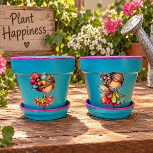 May include: Two bright blue ceramic plant pots with matching saucers, each featuring a painted illustration of a character with brown hair and a colorful dress. The rims of the pots and the edges of the saucers are painted in a vibrant pink color.