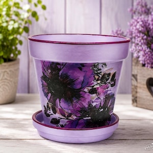 May include: A lavender-colored ceramic flower pot with a matching saucer, featuring a dark purple and black floral print on the front. The rim of the pot and the edge of the saucer are painted in a deep, solid purple, creating a distinct contrast.