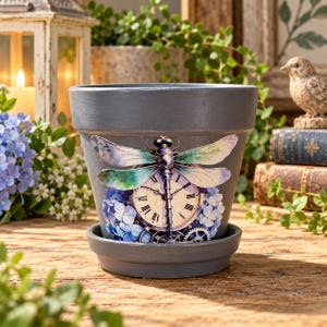 May include: A matte gray ceramic flower pot with a matching saucer, featuring a central graphic of a dragonfly with iridescent wings positioned over a vintage clock face and blue hydrangea flowers, set against a neutral gray background.