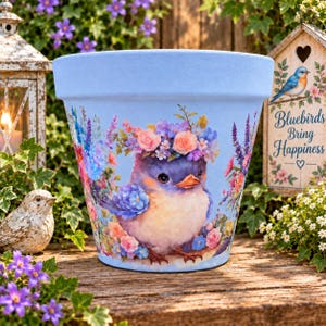 May include: A light blue flower pot featuring a central illustration of a small bird with a floral crown, surrounded by colorful wildflowers. The design includes soft pink, purple, and blue floral accents, creating a detailed botanical scene.