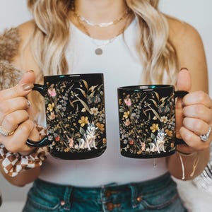 May include: Two black ceramic mugs featuring a floral design and a wolf illustration. The mugs are filled with a dark liquid.