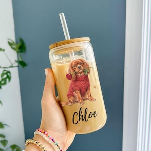 Peut inclure: Un gobelet en verre transparent avec un couvercle en bambou et une paille transparente. Le gobelet présente un dessin d'un Cavalier King Charles Spaniel portant un pull rouge et tenant une rose rouge. Le nom "Chloe" est écrit en cursive.