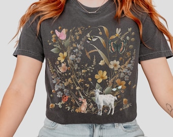 May include: Dark gray t-shirt featuring a whimsical floral design with butterflies, bees, a goat, and a rabbit. The design includes yellow and white flowers, and a ladybug. The shirt has short sleeves and a crew neck.