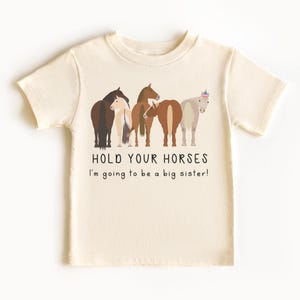 May include: A light beige t-shirt with a line of five horses, one wearing a party hat. The text "Hold your horses I'm going to be a big sister!" is printed below the horses.