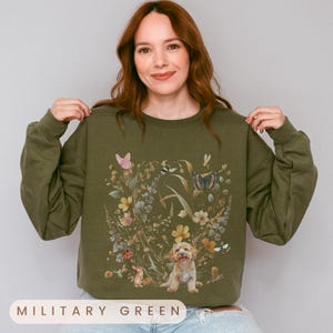 May include: A green sweatshirt with a floral print featuring a dog, butterflies, and other insects. The sweatshirt has a crew neck and long sleeves.