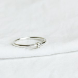 May include: A silver ring with a knot design. The ring is made of a shiny, metallic material and features a simple, elegant style. The knot detail is centered on the ring's band. The ring is shown on a white, textured surface.