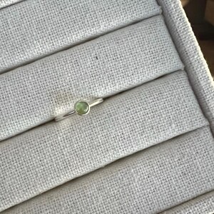 May include: A delicate silver ring featuring a small, round, pale green gemstone. The ring is set on a light gray, textured surface, showcasing the minimalist design and the gemstone's subtle color. The ring is a piece of jewelry.