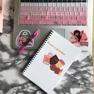 Könnte beinhalten: Ein Laptop mit rosa Tastaturabdeckung und Aufklebern, darunter einer mit der Aufschrift "That's My Purse! I Don't Know You!" und ein weiterer mit der Aufschrift "Latinas Uprising". Ein Spiralnotizbuch mit den Worten "Ponte a estudiar" und verschiedenen Illustrationen ist ebenfalls vorhanden.