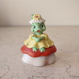 May include: A small ceramic figurine of a green frog wearing a yellow dress and a white flower crown, sitting on a red and white mushroom.