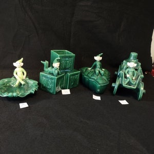 May include: Four green ceramic elf figurines. One elf is sitting on a leaf-shaped dish, another is sitting on a block with the letter "B" on it, another is sitting on a leaf-shaped dish, and the last elf is sitting on a green wagon.