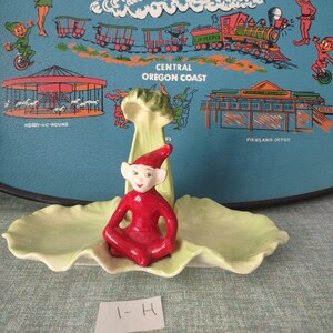 May include: A green ceramic dish shaped like a leaf with a red elf figurine sitting on top. The elf is wearing a red hat and has a white face. The dish has the text "Pixieland Depot" on it.