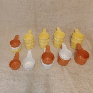 May include: A set of 12 vintage measuring cups in yellow, white, and orange. The cups are made of plastic and have a handle. They are arranged in three rows of four.