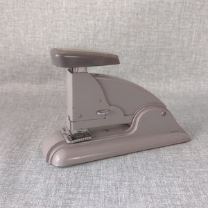 May include: A gray metal stapler with a silver lever and a black base. The stapler is sitting on a gray textured surface.