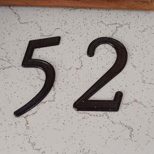 May include: Dark brown metal house numbers, the numbers 5 and 2, are displayed against a speckled white background. The numbers have a textured surface and are designed in a modern font.
