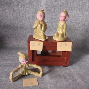 May include: Three ceramic elf figurines in shades of green and pink. Two are seated on a small wooden cabinet, and one is posed with arms and legs outstretched. Each figurine has a pointed hat and a smiling face. The tags read 2-A, 2-B, and 2-C.