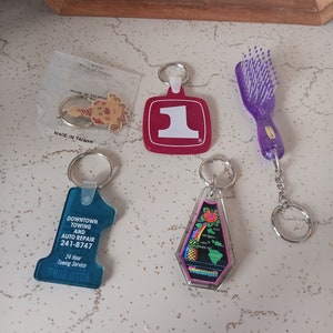 May include: Five keychains with different designs. A purple hairbrush keychain, a blue keychain with the text "Downtown Towing and Auto Repair 241-8747 24 Hour Towing Service", a red keychain with the number "1", a keychain with a cartoon clown, and a clear keychain with a map of Hawaii and the text "Hawaii".