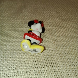 May include: A small ceramic figurine of Minnie Mouse wearing a red and white dress. The figurine is standing on a yellow base with the text "Bone China Disney Taiwan" printed on it.