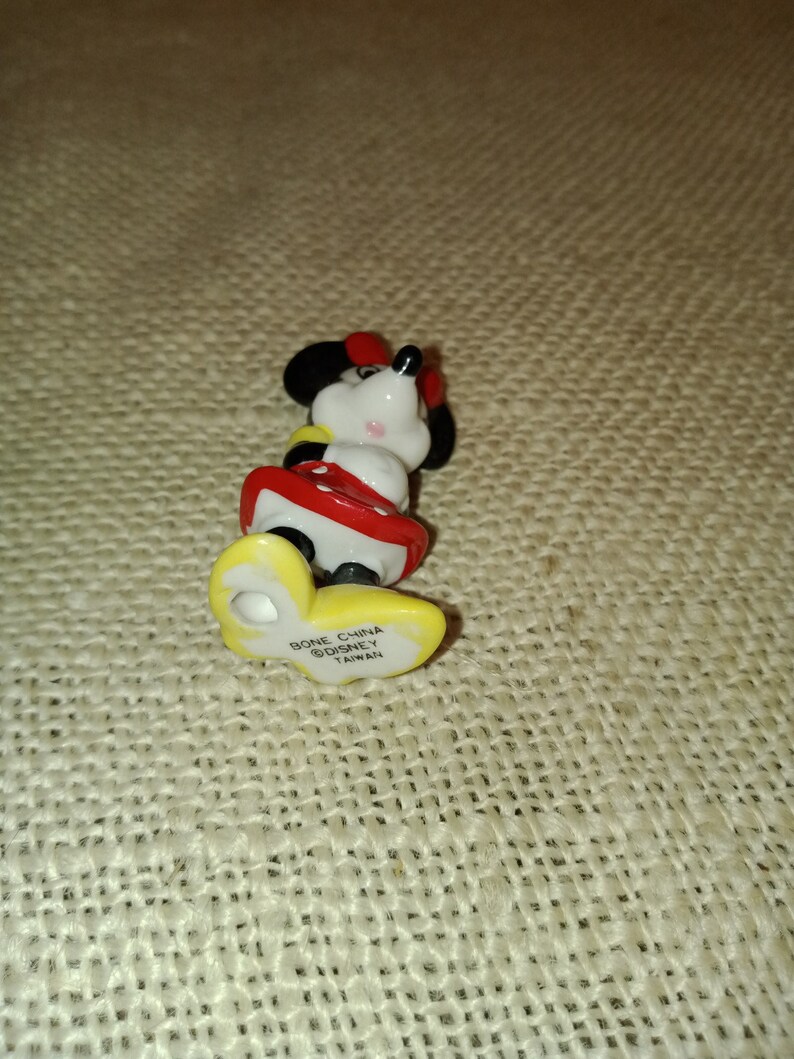 May include: A small ceramic figurine of Minnie Mouse wearing a red and white dress. The figurine is standing on a yellow base with the text "Bone China Disney Taiwan" printed on it.