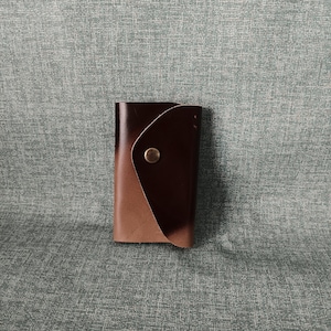 May include: A brown leather key holder with a flap closure and a metal button. The key holder is folded in half and is sitting on a textured surface.