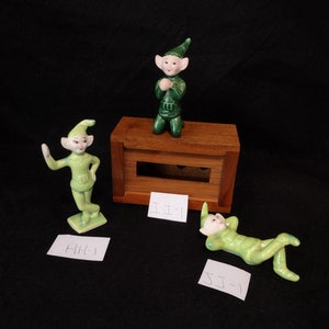 May include: Three green ceramic elf figurines, one kneeling, one standing with an arm raised, and one lying down. The figurines are on a black surface with handwritten labels.