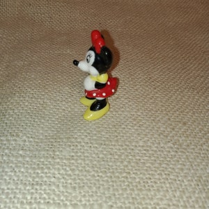 May include: A small ceramic figurine of Minnie Mouse. She is wearing a red dress with white polka dots, yellow shoes, and a red bow.