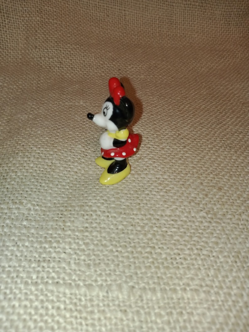 May include: A small ceramic figurine of Minnie Mouse. She is wearing a red dress with white polka dots, yellow shoes, and a red bow.