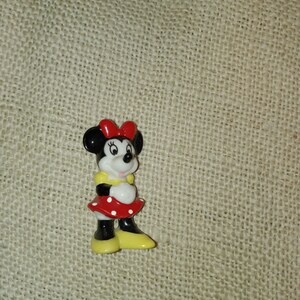 May include: A small ceramic figurine of Minnie Mouse. She is wearing a red dress with white polka dots, a yellow bow, and yellow shoes.