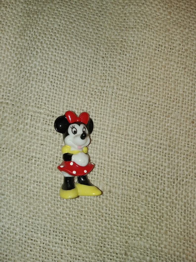 May include: A small ceramic figurine of Minnie Mouse. She is wearing a red dress with white polka dots, a yellow bow, and yellow shoes.