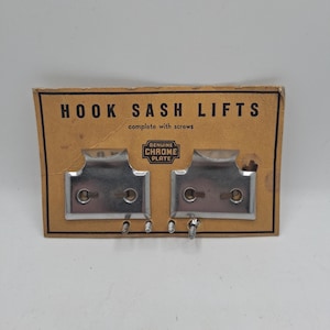 May include: A package of two chrome-plated hook sash lifts with screws. The cardboard packaging is brown with black text that reads "HOOK SASH LIFTS" and "complete with screws". The package also has a "GENUINE CHROME PLATE" emblem.