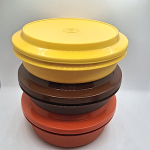 May include: A stack of four vintage plastic food storage containers in yellow, brown, and orange. Each container has a round base and a matching lid. The containers are stacked on top of each other.