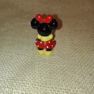 May include: A small ceramic figurine of Minnie Mouse. She is wearing a red dress with white polka dots and a red bow. Her ears are black and her shoes are yellow.