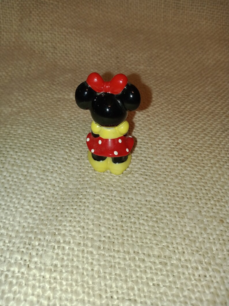 May include: A small ceramic figurine of Minnie Mouse. She is wearing a red dress with white polka dots and a red bow. Her ears are black and her shoes are yellow.