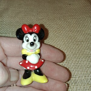 May include: A small ceramic figurine of Minnie Mouse. She is wearing a red dress with white polka dots, a yellow shirt, and a red bow. Her ears are black and her face is white with black eyes and a pink nose.