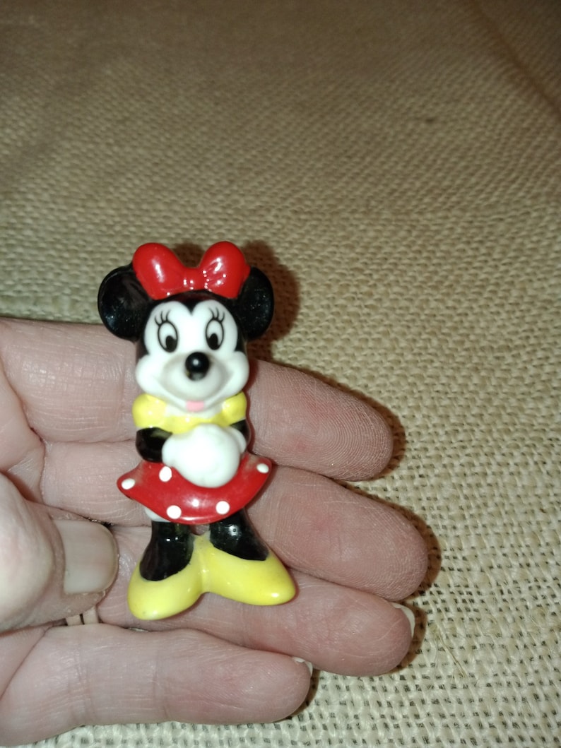 May include: A small ceramic figurine of Minnie Mouse. She is wearing a red dress with white polka dots, a yellow shirt, and a red bow. Her ears are black and her face is white with black eyes and a pink nose.