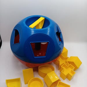 May include: A blue and red sphere-shaped toy with various cut-out shapes. Includes yellow geometric blocks in shapes like a star, cross, and cylinder. A yellow handle is inserted into the top. Educational toy for toddlers.