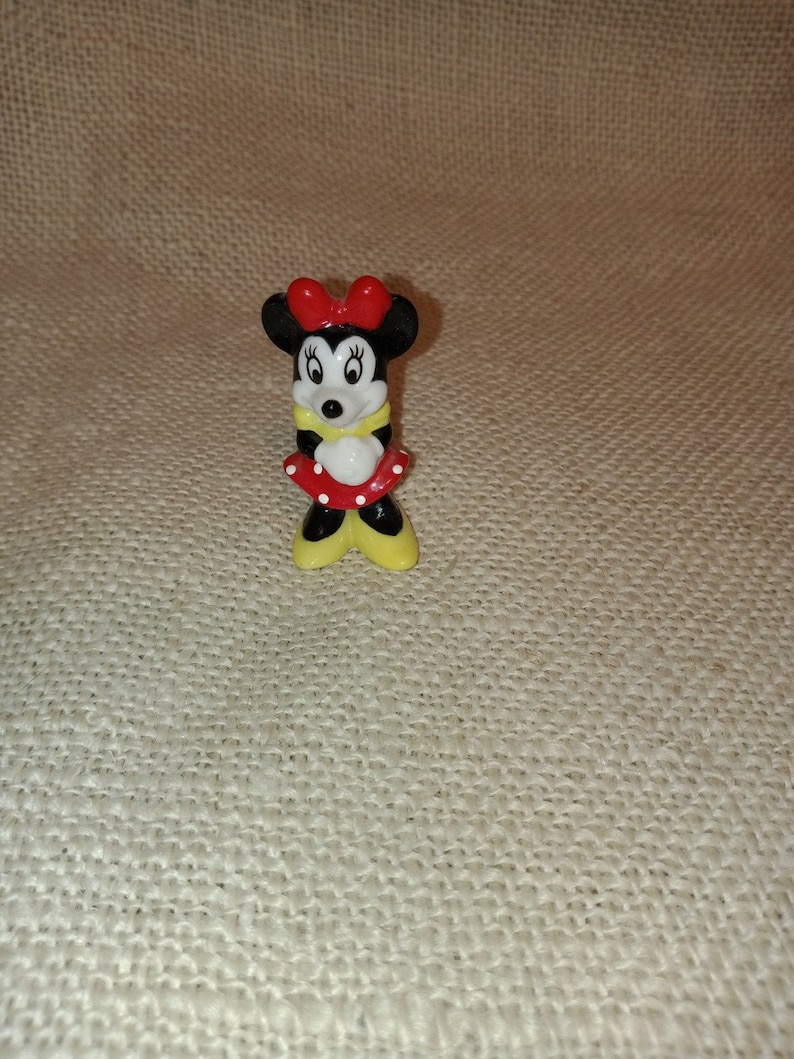 May include: A small ceramic figurine of Minnie Mouse. She is wearing a red dress with white polka dots, a yellow shirt, and a red bow. Her eyes are black and her nose is pink.