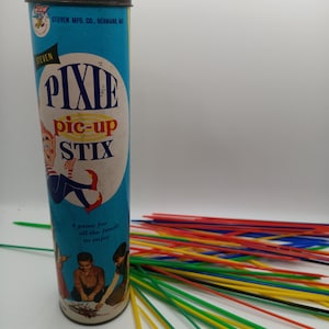 May include: A vintage "Pixie Pic-Up Stix" game set. The cylindrical container is light blue with the game's name and a cartoon illustration. Colorful sticks in various colors are scattered around the container. The text on the container reads "A game for all the family to enjoy."