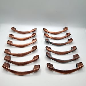 May include: A set of twelve curved drawer pulls arranged in two columns. The pulls feature a metallic copper finish with some showing darker, oxidized patina accents along the center and edges of the arched handle design.