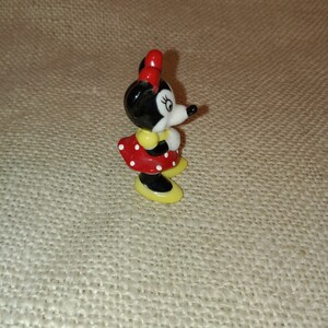 May include: A small ceramic figurine of Minnie Mouse. She is wearing a red dress with white polka dots, yellow shoes, and a red bow.
