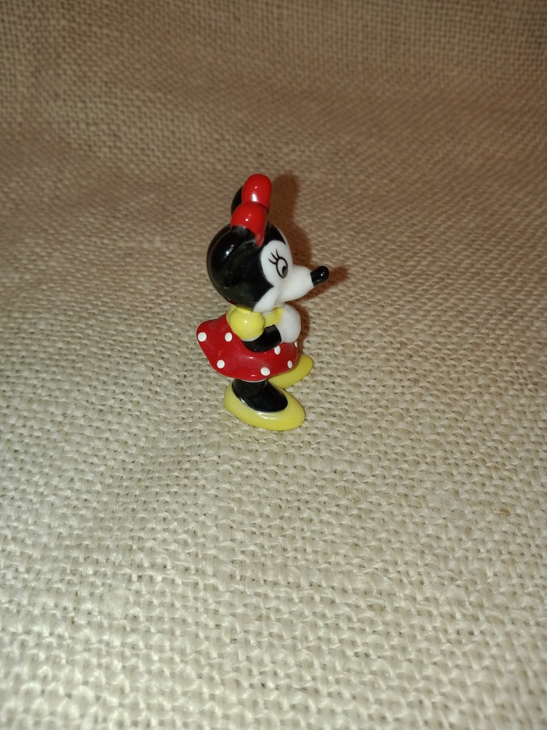 May include: A small ceramic figurine of Minnie Mouse. She is wearing a red dress with white polka dots, yellow shoes, and a red bow.